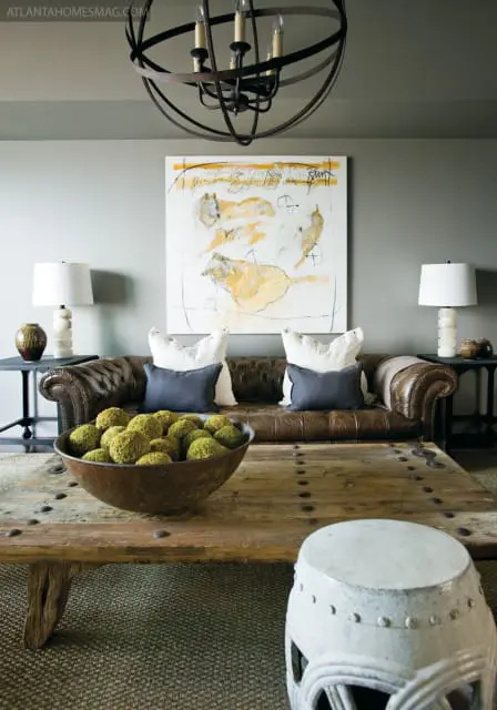Living room with brown leather Chesterfield sofa, abstract art, large wooden coffee table with bowl of moss balls, and globe chandelier.