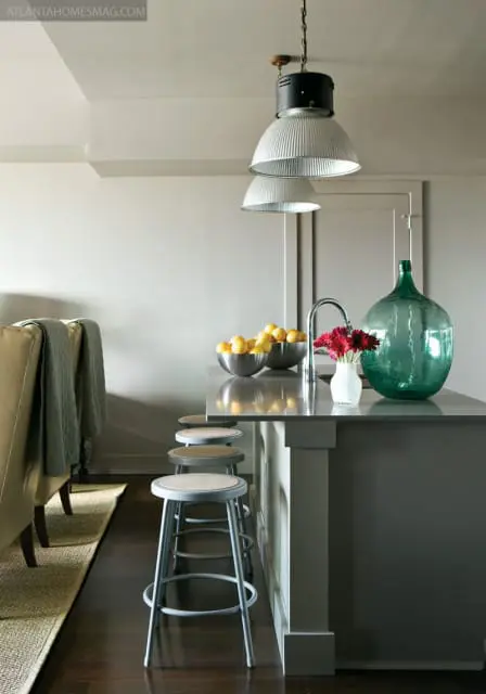 Gray kitchen island with stainless steel stools, bowls of lemons, a vase of red flowers, and a large green glass bottle.
