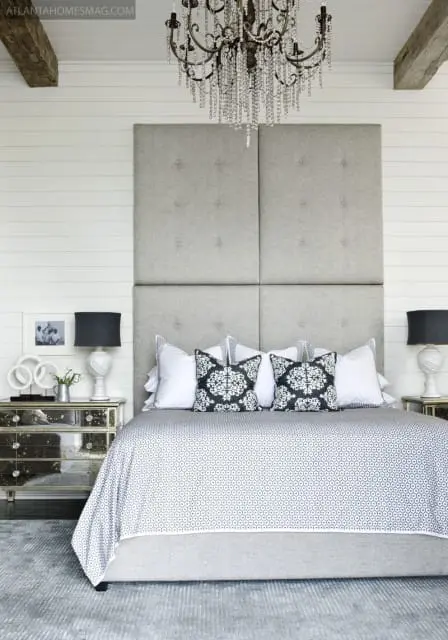 Bedroom with a large tufted grey headboard, white shiplap walls, a crystal chandelier, and a bed dressed in patterned bedding and black and white accent pillows.