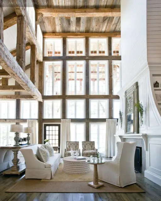 Great room with a vaulted ceiling featuring exposed wooden beams and rustic wood paneling, a wall of windows, a white sofa, two patterned armchairs, and a large woven coffee table.