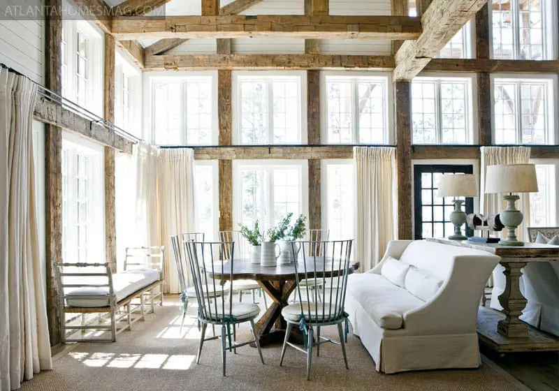 Rustic dining room with exposed wooden beams, large windows with white curtains, a round wooden table with metal chairs, and a white sofa.