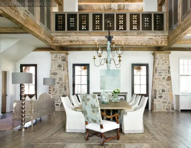 Dining room with rustic wooden beams, stone fireplace, white shiplap walls, and a long wooden table with slipcovered chairs.