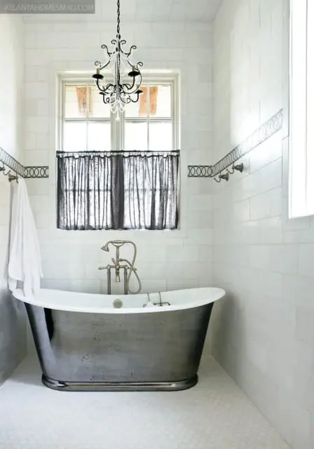 White subway tile bathroom with a dark metal freestanding tub, hexagonal floor tiles, a black chandelier, and a window with dark linen curtains.