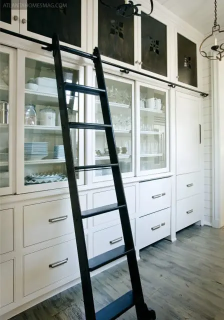 White built-in cabinetry with glass doors displaying dishes and glassware, topped with dark wood panels featuring cut-out designs. A black rolling ladder leans against the cabinets.