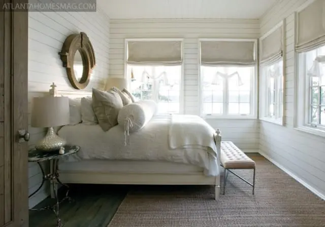 Bedroom with white shiplap walls, a white bed with neutral linens, a round wooden mirror, and windows with linen roman shades.