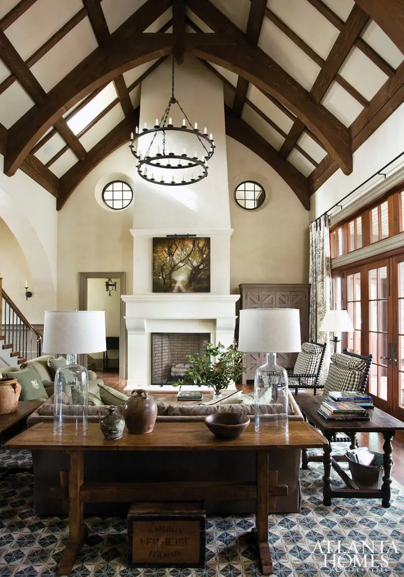 Living room with vaulted wood-beamed ceiling, stone fireplace with artwork, two large lamps on a coffee table, and patterned rug.