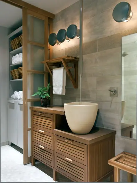 Bathroom with wood vanity featuring slatted drawers, a stone vessel sink, and a wooden shelf with a towel. A sliding glass door reveals a closet with stacked towels and baskets.