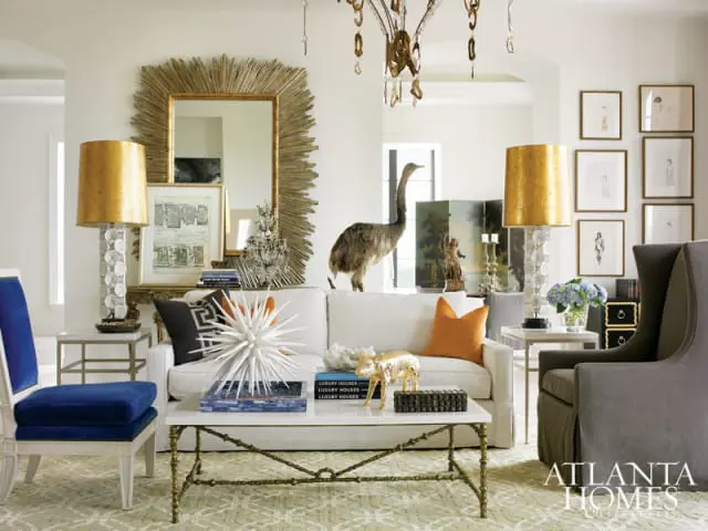 Living room with white sofa, blue velvet chair, gold sunburst mirror, and eclectic decor including a taxidermy bird and gold fox figurine.