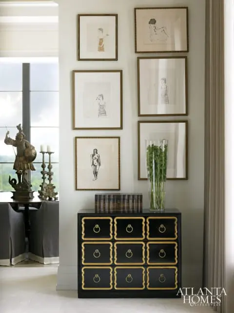 Black dresser with gold trim and ring pulls, topped with a wooden box and a glass vase of greenery, beneath a gallery wall of framed figure drawings.