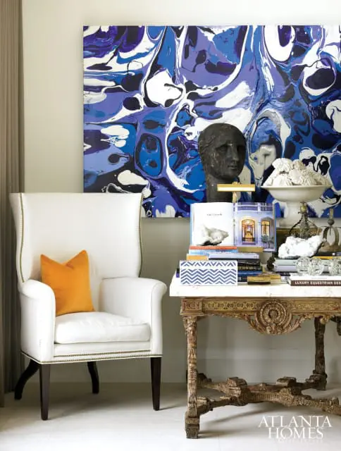 White wingback chair with orange pillow next to a marble-top table with ornate carved base, displaying books and decorative objects under abstract blue and white artwork.