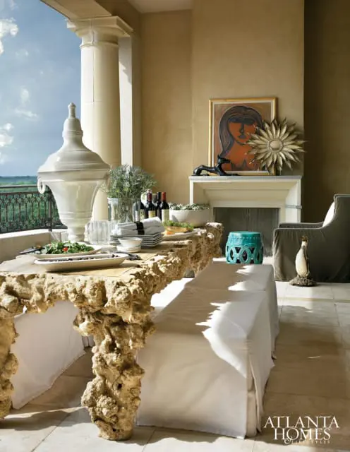 Covered patio with a rough-hewn stone dining table set for a meal, a fireplace with artwork, and a view of the landscape beyond a balcony