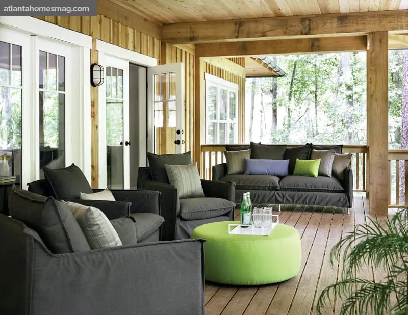 Covered porch with dark gray upholstered seating, a bright green ottoman coffee table, and French doors opening to a wooded area.
