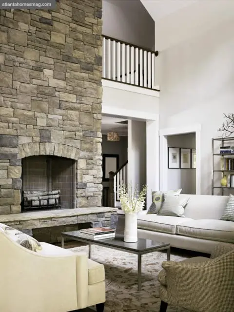 Living room with a large stone fireplace, neutral-toned sofa and armchairs, and a dark coffee table with a vase of white flowers.