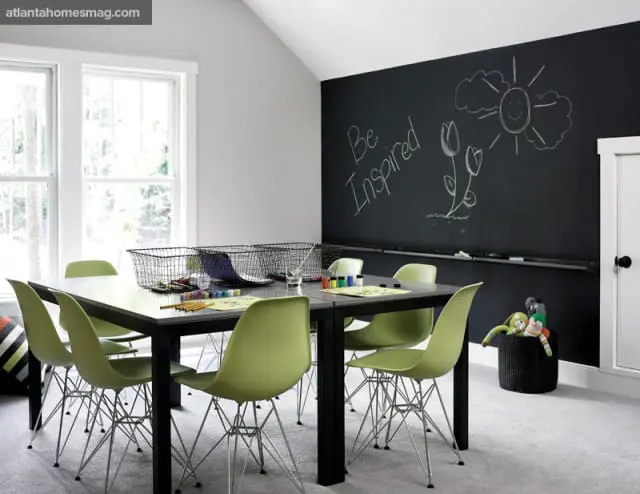 Playroom with a large black chalkboard wall featuring chalk drawings, a dark rectangular table with lime green chairs, and wire baskets on the table.