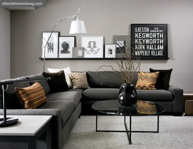 Gray sectional sofa with patterned throw pillows and a black vase with branches on a glass coffee table. A shelf with framed art hangs on the wall above the sofa.
