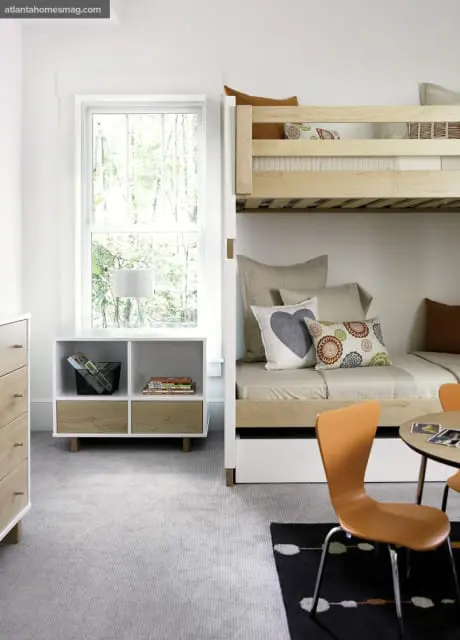 Children's bedroom with light wood bunk beds, white storage unit with wood drawers, and orange chairs at a small round table.