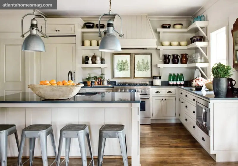 Cream kitchen with dark countertops, stainless steel stools, a large wooden bowl of oranges, and two framed pine tree prints above the stove.
