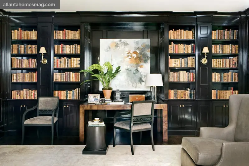 Dark wood library with floor-to-ceiling built-in bookshelves filled with books, a wooden desk with artwork and a lamp, and upholstered chairs.