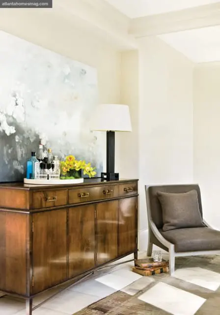 Wooden sideboard with brass hardware displaying bottles and yellow flowers, next to a chair and abstract art.