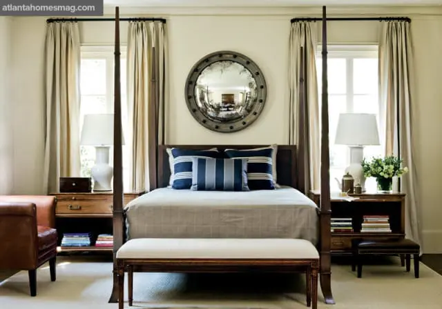Bedroom with dark wood four-poster bed, neutral bedding, navy and white striped pillows, a round distressed mirror, and tan curtains.