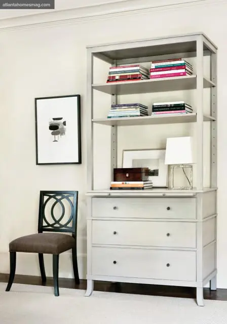 Light gray open hutch with three drawers below, displaying books, a wooden box, and a small lamp. A black chair with a patterned seat sits to the left.