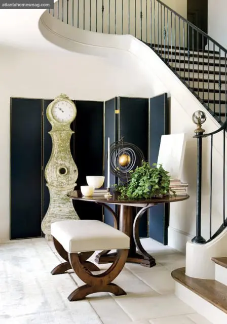 Entryway with a tall, distressed green Mora clock, a dark folding screen, a round wooden table with decorative objects, and a cream upholstered bench. A curved staircase with a black railing is visible.