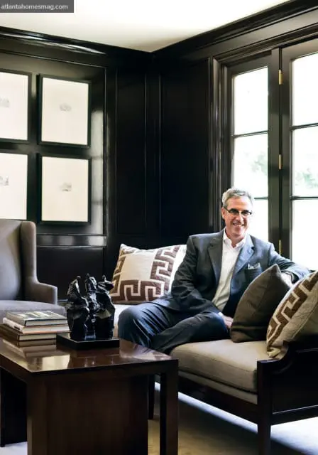 Man in gray suit sitting on a sofa in a room with dark paneled walls, framed artwork, and a dark wood coffee table with sculptures.