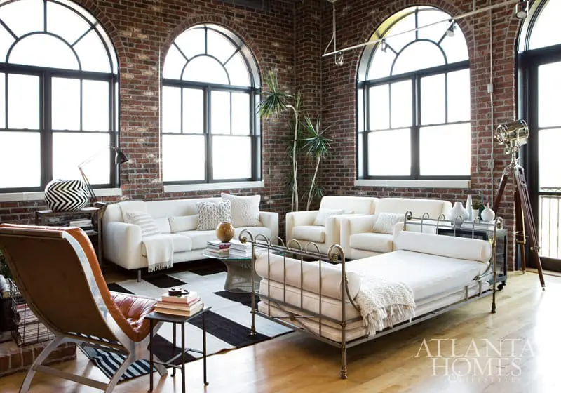 Loft living room with exposed brick walls, arched windows, white sofas, a metal daybed, and a black and white striped rug.