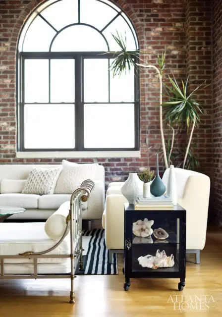 Loft living room with exposed brick walls, a large arched window, cream sofas, a metal daybed, and a black side table displaying crystals and vases.