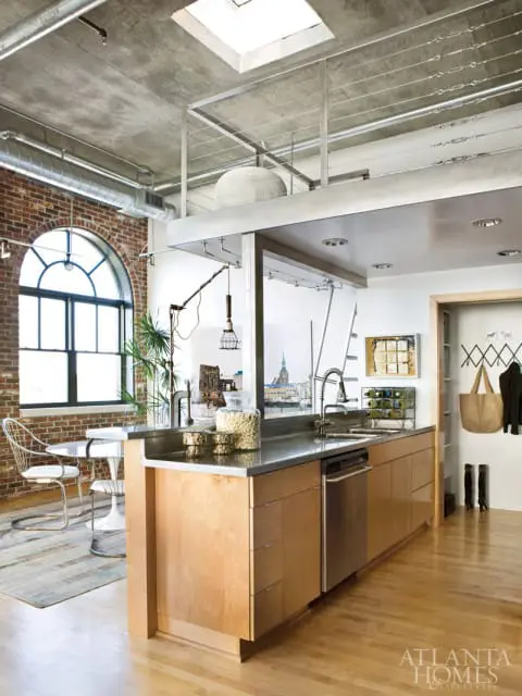 Loft kitchen with exposed brick wall, arched window, concrete ceiling, metal railings, and light wood cabinetry. A stainless steel island with a sink separates the kitchen from a dining area.
