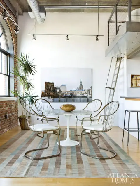 Loft dining area with a marble tulip table, chrome chairs, and a large cityscape artwork above a striped rug. Exposed brick and industrial elements are visible.