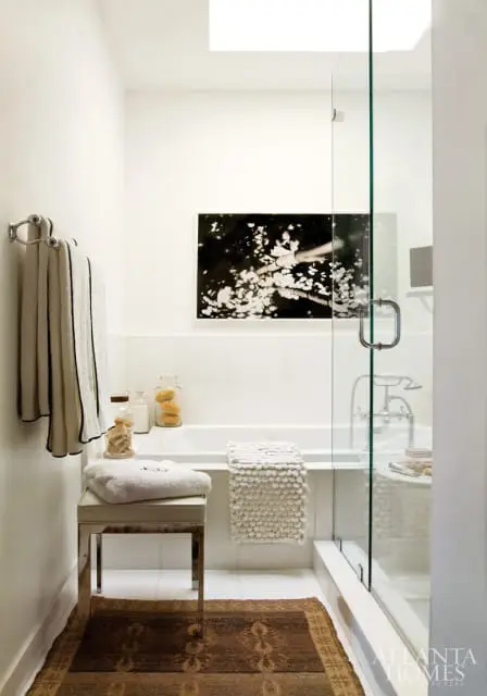White bathroom with a glass-enclosed shower, bathtub, towel rack with striped towels, a stool with towels, and a dark patterned rug on the floor.