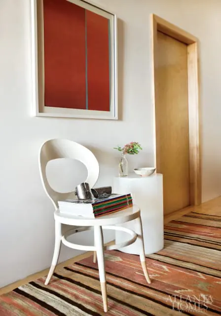 White chair with stack of magazines and silver bracelets, next to a white pedestal with a vase and bowl. Abstract red art hangs on the wall above.