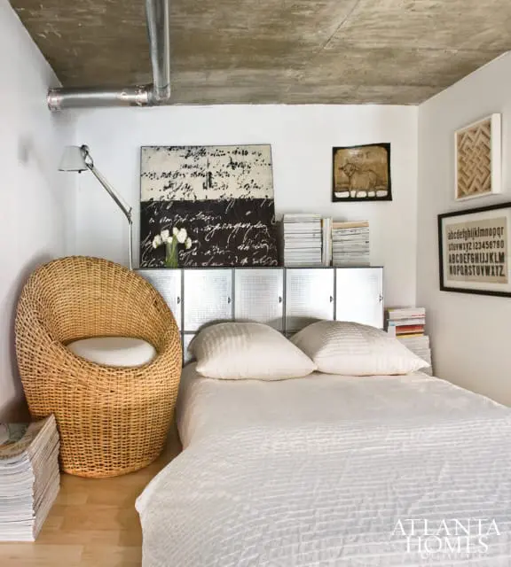 Bedroom with a large wicker chair, a bed with white bedding, and a metal cabinet headboard. The ceiling is concrete with exposed pipes.