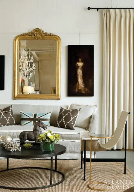 Living room with a light gray sofa adorned with patterned pillows and a Union Jack pillow, a gilded ornate mirror, and a dark abstract painting of a woman in a gown.