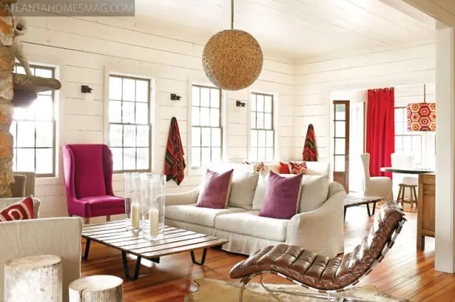 Living room with white shiplap walls, a cream sofa with purple pillows, a pink wingback chair, a wood slat coffee table, and a large woven pendant light.