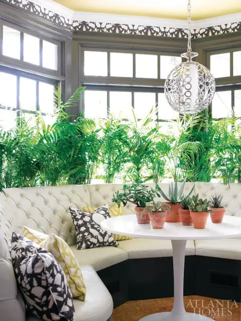 Tufted white banquette seating with patterned pillows surrounds a white tulip table with potted succulents. A decorative silver pendant light hangs above.