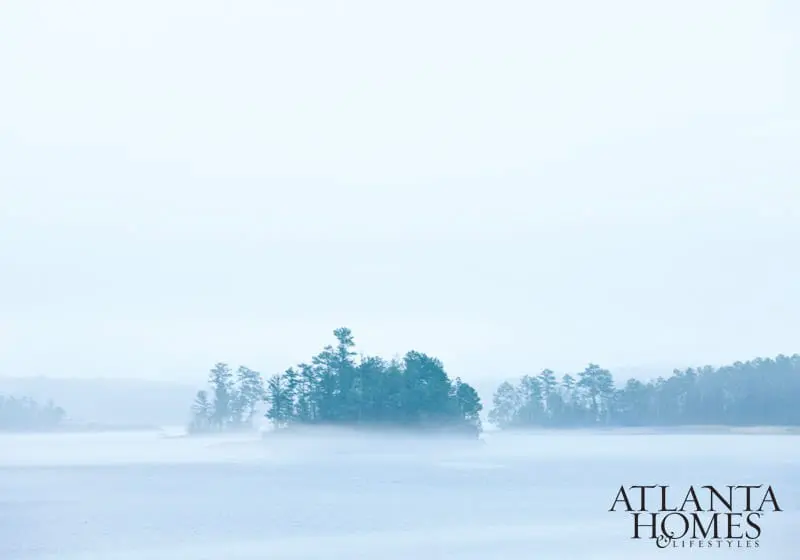 Misty lake with a small island covered in pine trees, and a forest in the background. Atlanta Homes & Lifestyles logo in the bottom right corner.