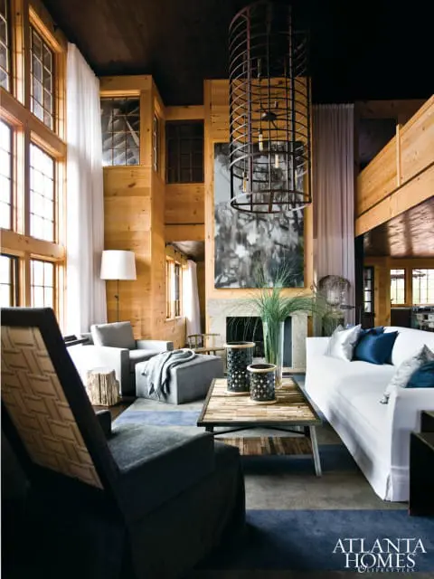 Living room with wood-paneled walls, large windows, a white sofa, gray armchairs, a rustic coffee table, and a large cage-like chandelier.