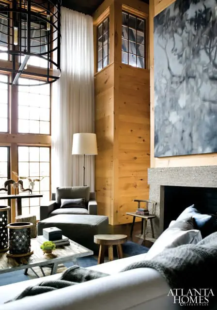 Living room with wood-paneled walls, large windows, a gray armchair and ottoman, a concrete fireplace, and abstract art above the mantel.