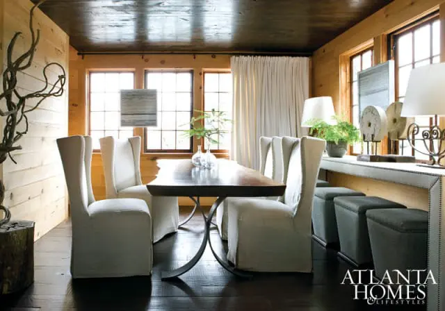 Dining room with wood-paneled walls, dark wood ceiling, and dark wood floors. Features a live-edge dining table with metal base, upholstered dining chairs, and a console table with lamps and decorative objects.