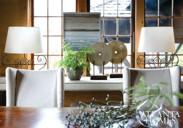Dining room with two upholstered chairs flanking a table, featuring decorative stone discs, a potted fern, and two lamps against a window