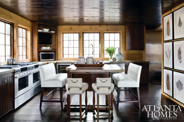 Rustic kitchen with wood-paneled walls and ceiling, white double oven range, butcher block island with white bar stools, and framed botanical prints.