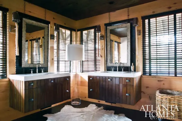 Rustic bathroom with dual wood vanities topped with white countertops and undermount sinks, each with a textured frame mirror above. Wood-paneled walls and dark blinds adorn the windows.