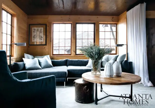 Living room with wood-paneled walls and ceiling, featuring a dark blue sectional sofa, light blue throw pillows, a round wooden coffee table with decorative vases, and a tall floor lamp.
