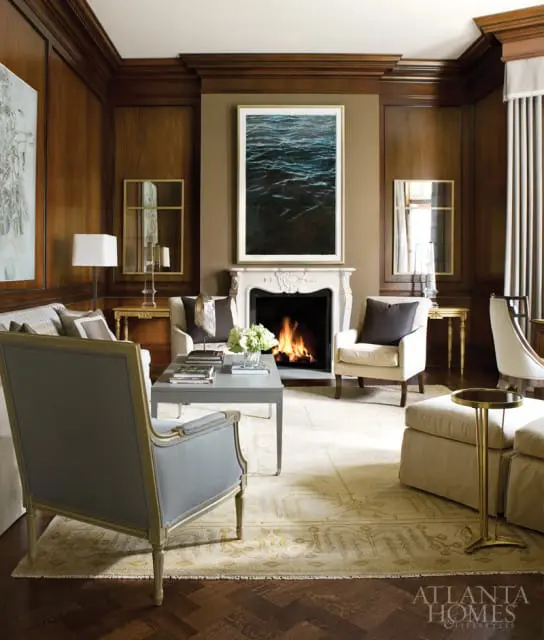 Living room with dark wood paneled walls, a white marble fireplace with a large artwork of dark water above, and upholstered furniture.