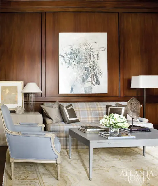 Living room with dark wood paneled walls, a plaid sofa, a light blue armchair, and a grey coffee table. Abstract art hangs above the sofa.