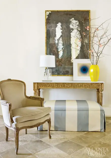 Entryway with a cream upholstered armchair, a console table with a gold ornate frame, and a striped ottoman in cream and blue.