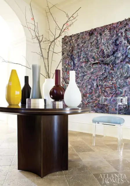 Entryway with a dark wood pedestal table holding various colored vases and branches, a textured purple artwork on the wall, and a clear acrylic stool with a blue cushion.