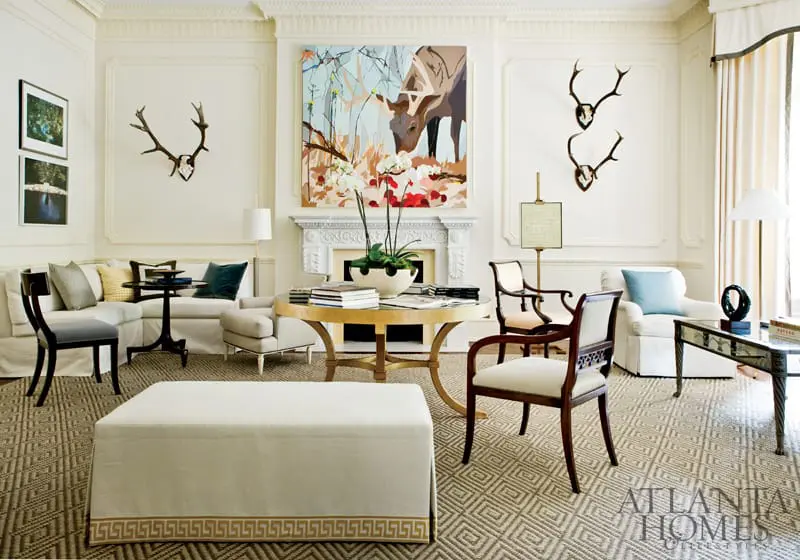Living room with a sectional sofa, upholstered ottoman, round dining table, fireplace, and artwork. The walls are white with ornate molding.